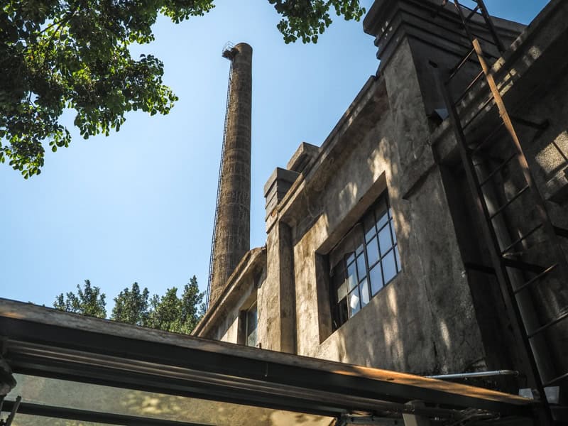 A smokestack riding up from behind some old factory buildings and trees