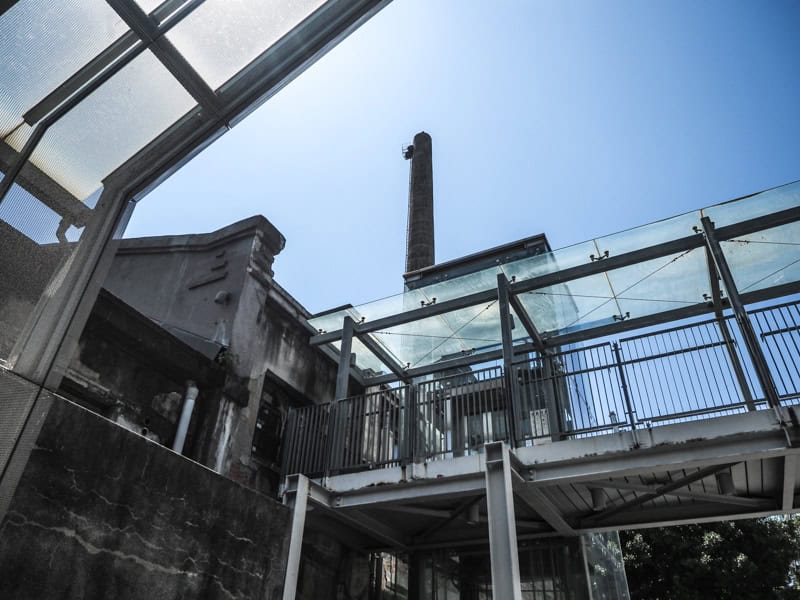 Looking up past some glass and metal elevated walkways and old cement buildings at a blue sky pieced by towering smokestack