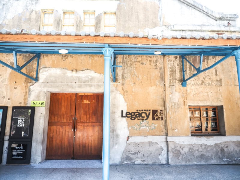 Exterior of an old beige building with wooden door and a sign that says Legacy in black