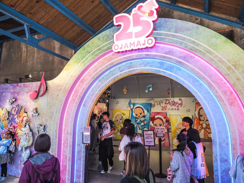 An arched entranceway with blue, pink, and green rainbow, people below, and sign indicating it is an Ojamajo Doremi 25 year anniversary pop-up