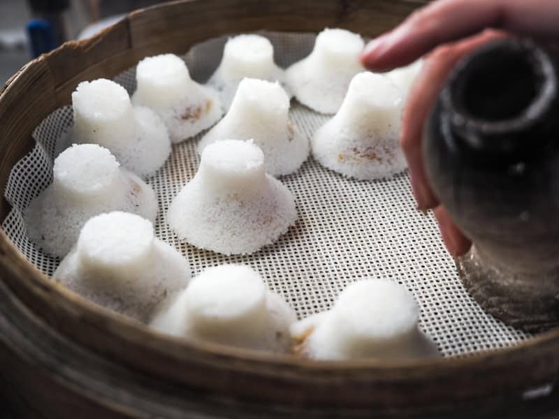 A large round bamboo basket with a dozen white hat-shaped scholar's cakes on it and a cat pressing one more down out of a mold