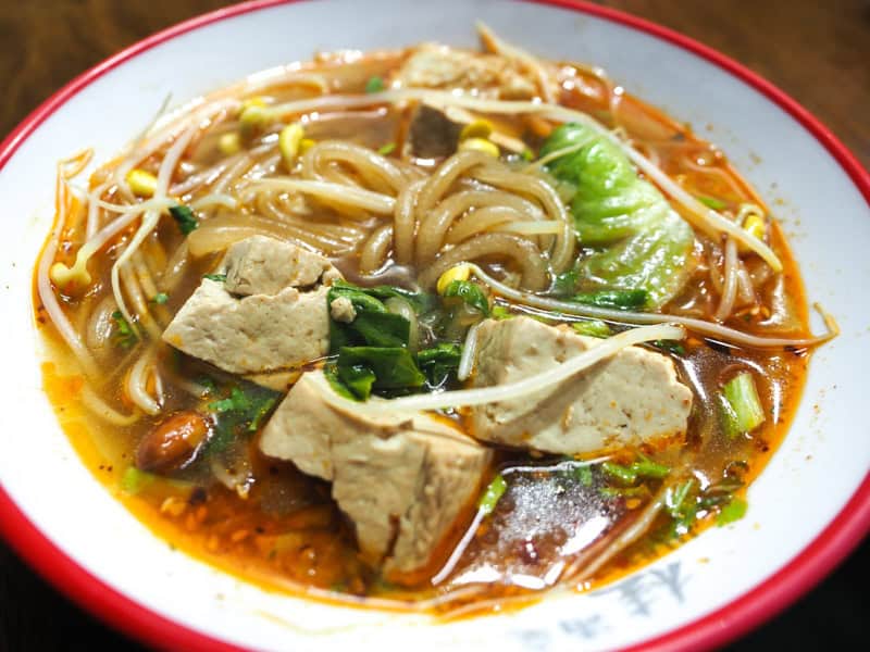 Close up of a bowl of spicy noodle soup with tofu, greens, peanuts, and bean sprouts