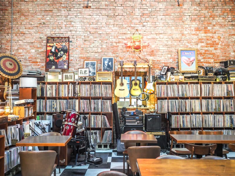 Some tables in a bar with shelves of records lining a red brick wall and some musical instruments at the back