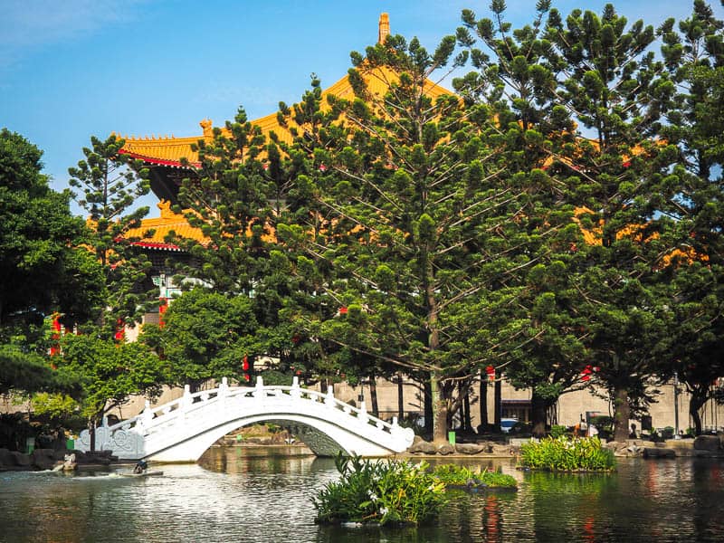 A pond with white arched bridge over it and orange roofed Chinese building behind the trees behind it