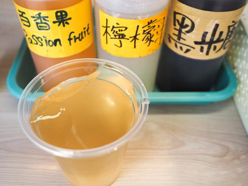 A plastic cup of aiyu jelly on a table with three containers of syrup behind