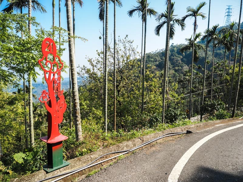 A curve in a road with red sign that has the number 21, palm trees lining the road, and lush mountain view beyond