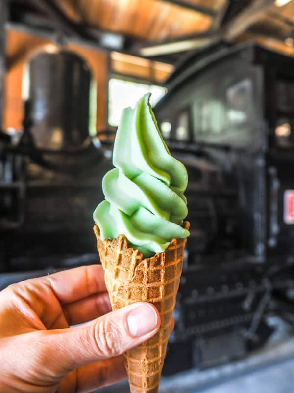 A hand holding up a green wasabi flavored soft serve ice cream on waffle cone in front of a black locomotive of the Alishan Forest Railway