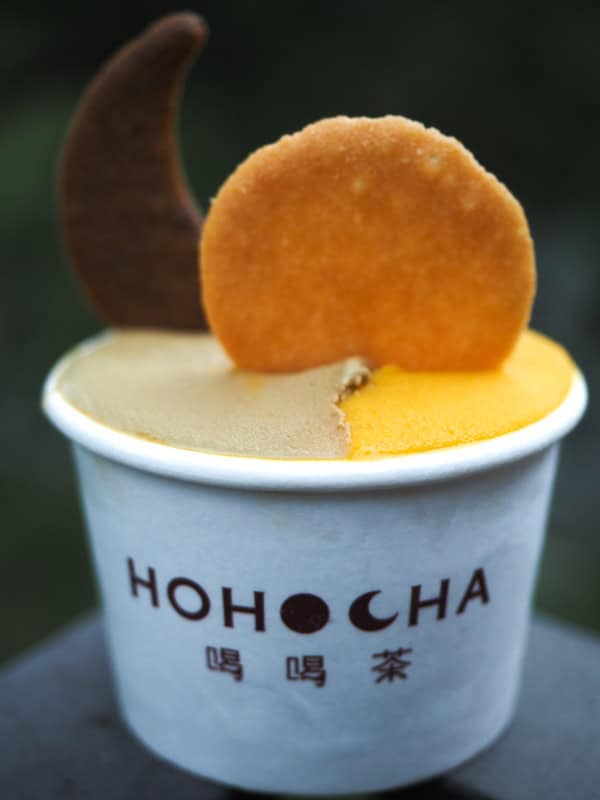 A small white Hohocha-branded paper cup with half orange and half brown gelato and two cookies sticking out, shaped like a sun and moon, on the corner of a balcony railing