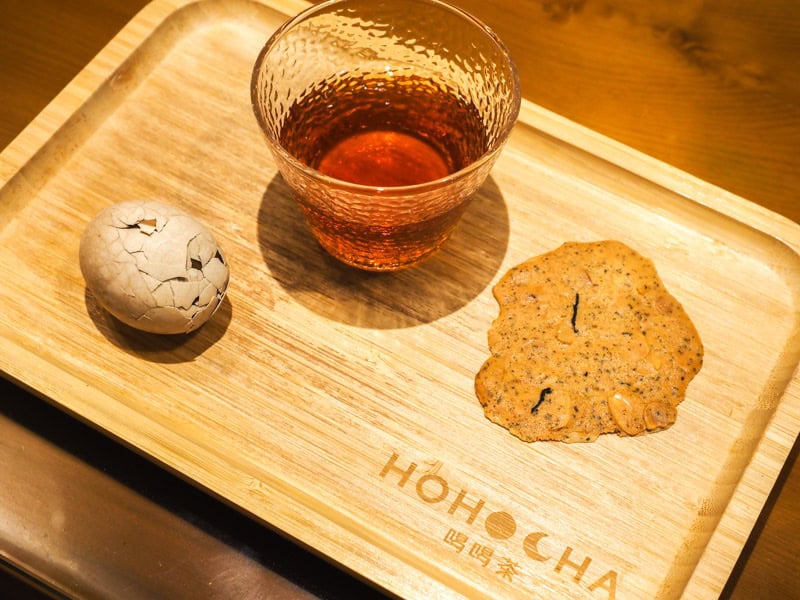 A wooden tray with the word Hohocha on it, a tea egg, a glass of black tea, and a cookie