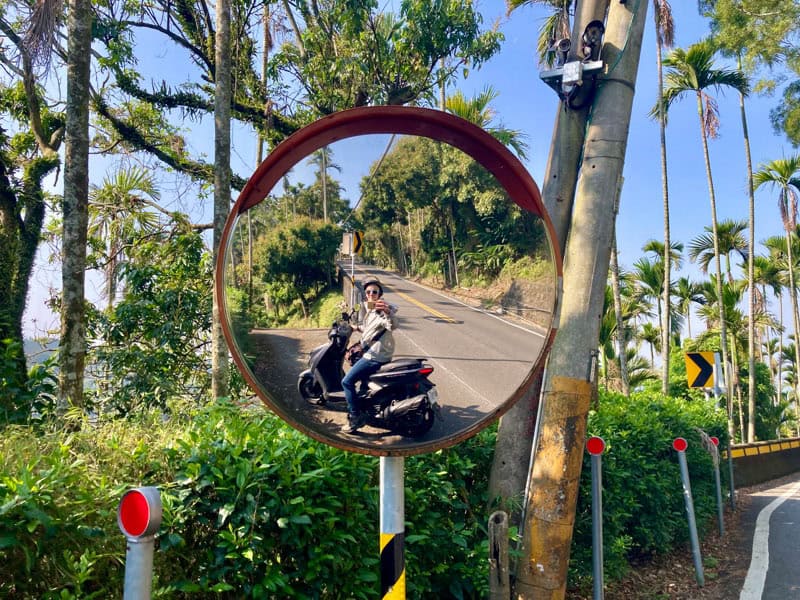 Nick Kembel taking a photo of himself in a round roadside mirror while sitting on a scooter on a small road in Chiayi