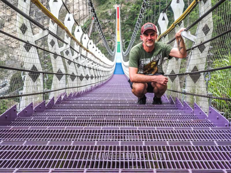 Nick Kembel kneeling down on the purple section of Shuiyuan Rainbow Suspension Bridge, with the other colors of the bridge visible in the distance behind him