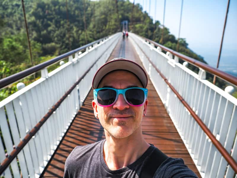 Selfie of Nick Kembel wearing cap and blue & pink sunglasses standing on Taiping Suspension Bridge