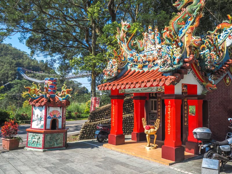 A small Taiwanese Taoist shrine with red columns and colorful, ornate roof, with large trees behind and part of Taiping Suspension Bridge visible below the branches
