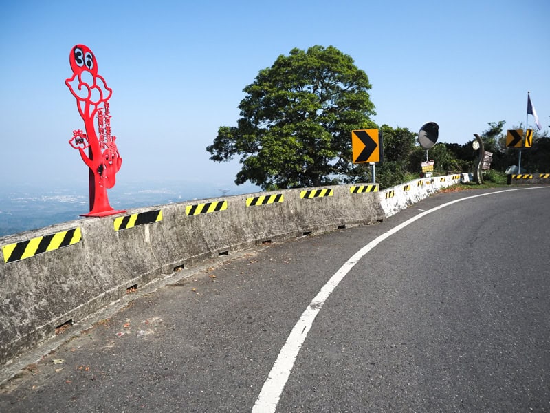 The curving side of a highway with cement rails, a red sign that says 36, and a few trees and blue sky beyond