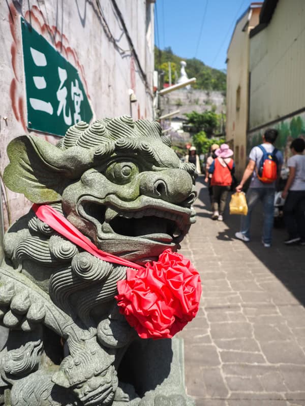 A stone lion at the side of a narrow alley with a red bow around its neck