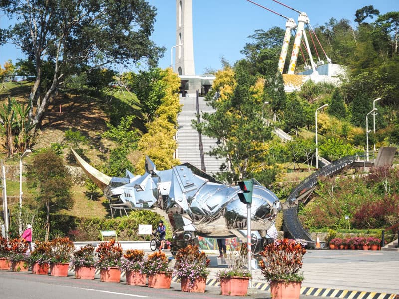 Looking across a road lined with potted plants at a large, metallic, robot rhino, and behind it there are stairs leading up to the entrance to Taiping Suspension Bridge