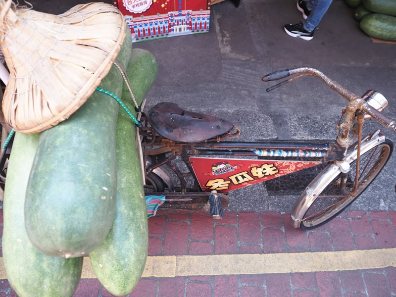 An old rusty bike with three huge winter melons tied to the back and topped with a conical farmer's hat