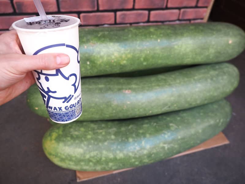 A hand holding a white paper cup of winter melon tea in front of a stack or enormous winter melons on the street