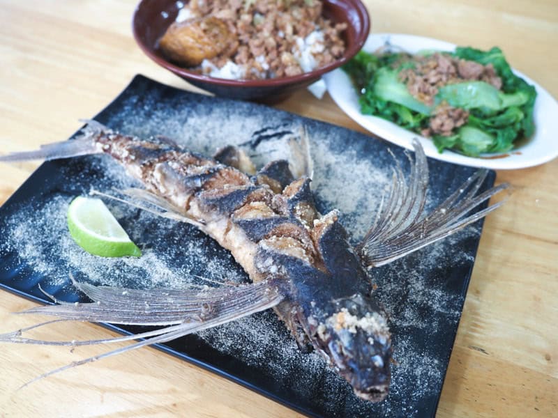 A whole deep fried flying fish with wings on a black plate with lime wedge, with a bowl of braised pork rice and small plate of green veggies behind