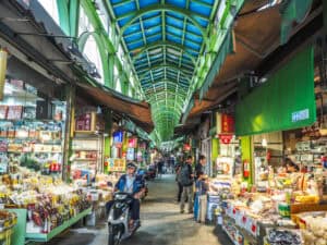 Looking down a long, covered traditional market street with green panels, a few customers browsing, and a man on scooter driving towards us