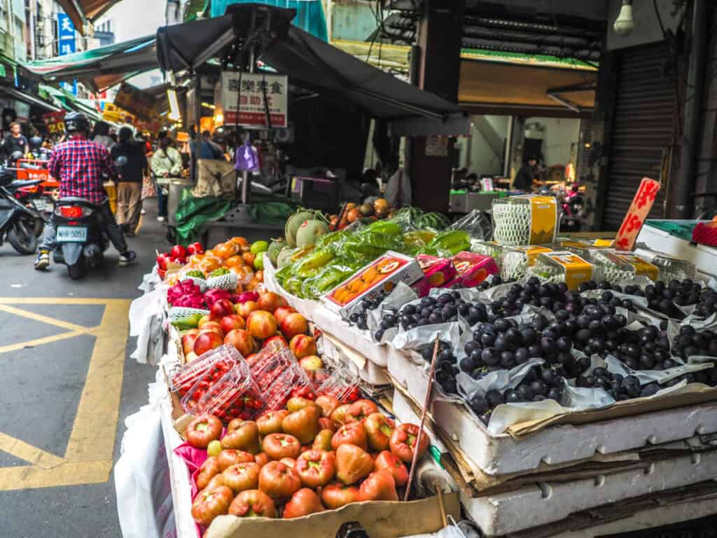A display of fruits in a market with narrow alley full of stalls and people past it