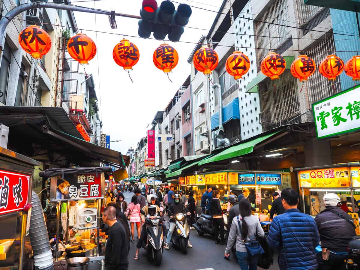 Scooters and pedestrians on a narrow lane lined with food stalls and with a string of red lanterns above