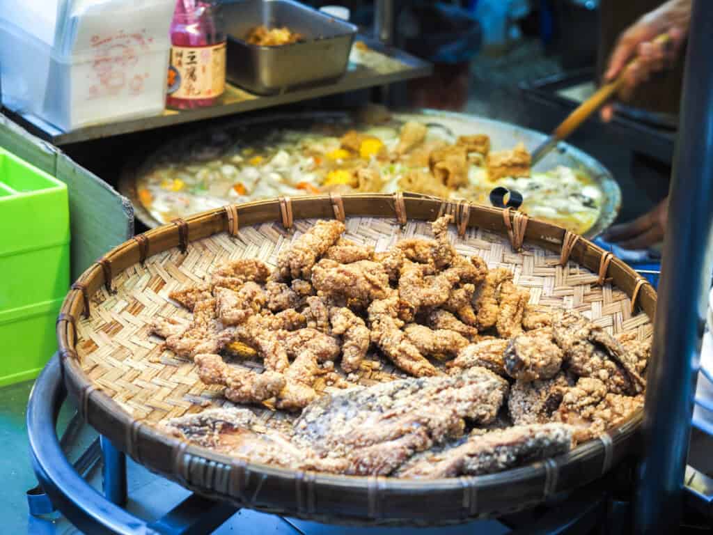 A large bamboo basket with pieces of deep fried fish and fish head on it, with large cauldron of cabbage and tofu stew behind being stirred by the vendor