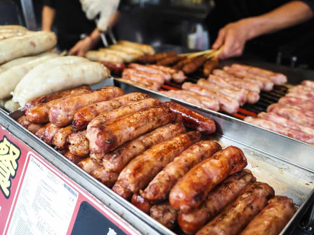 Close up of a tray of grilled sausages and grill with even more of them behind and vendor tending to them