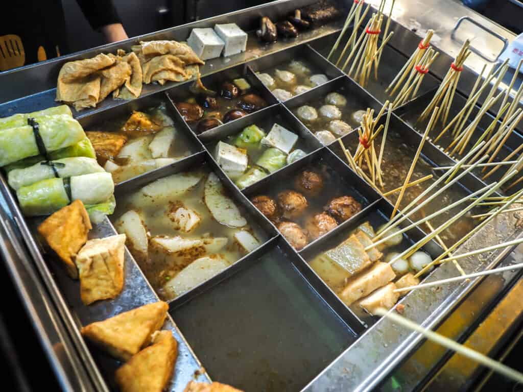 A large metal vat full of daishi broth and different foods on skewers in different compartments