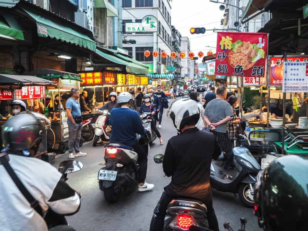 A busy scene in Sanmin Night Market with scooters and food vendor signs on the sides