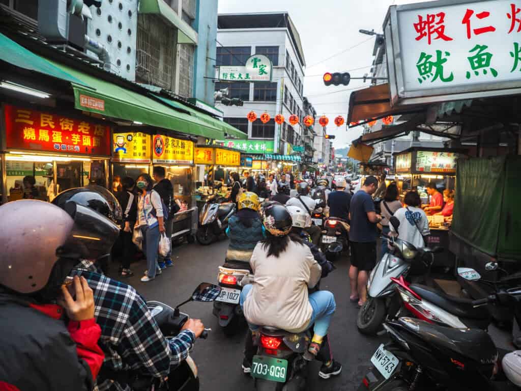 A narrow street packed with scooters and night market vendors on either side