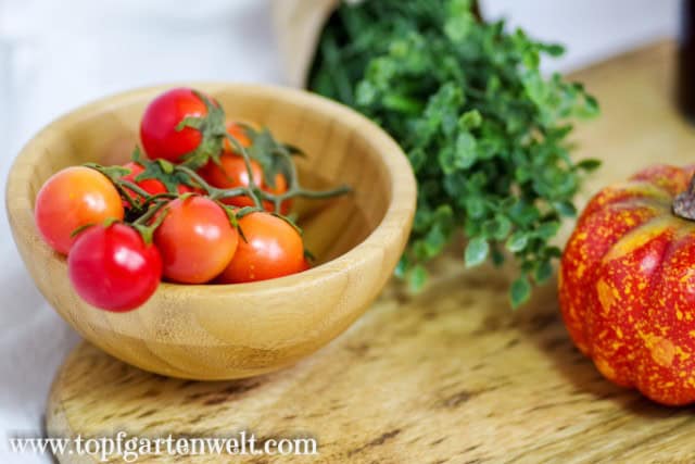 In einer Holzschüssel befindet sich eine Rispe mit Tomaten. Im Hintergrund sind Gewürze zu sehen.