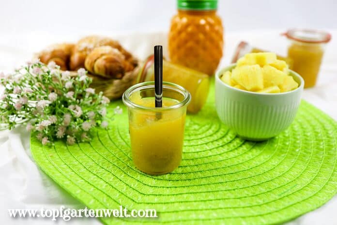 Ananasmarmelade in einem Weckglas. Im Hintergrund ist eine Schale mit Obst zu sehen.