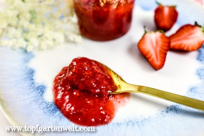 Ein Löffel mit Erdbeer-Holunderblüten-Marmelade liegt auf einem Frühstücksteller. Dahinter ist ein Glas mit dem Fruchtaufstrich zu sehen.