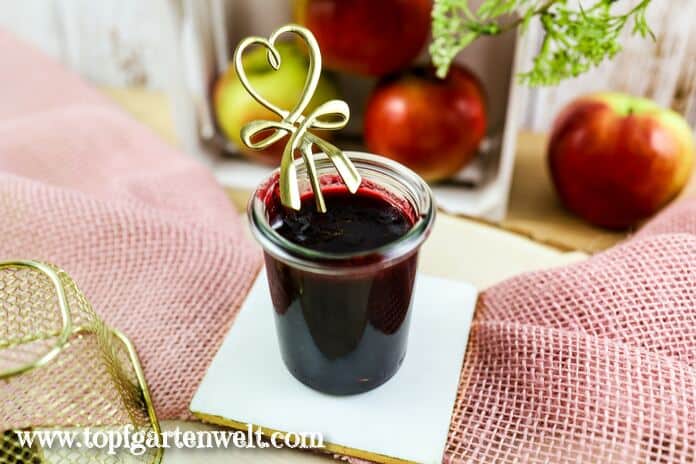 Ein kleines Weckgläschen gefüllt mit Apfel-Holunder-Marmelade steht auf einer weißen Fliese. Ein dekorativer Löffel ist bereits hergerichtet.