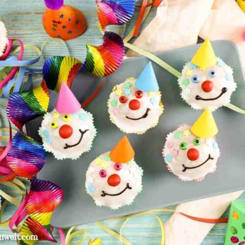 Fünf lustige Clown Muffins auf einem Teller. Jeder zeigt ein anderes Lächeln. Die Augen sind aus Smarties, die Haare bestehen aus Zuckerstreusel. Im Hintergrund befinden sich bunte Hütchen und Papierschlangen.