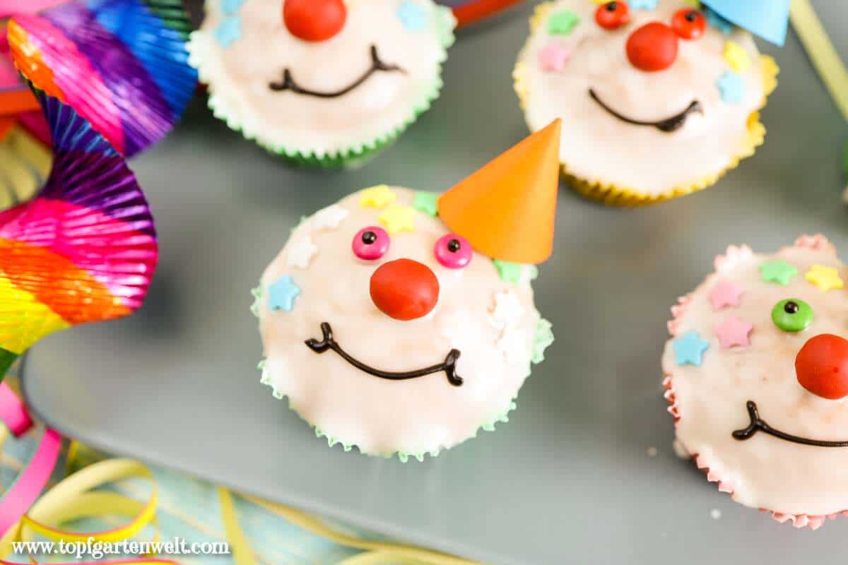 Auf dem Bild sind lustige Clown Muffins mit Gesicht und Hütchen zu sehen. Die Augen sind bunt. Die Nase ist rot. Sie lächeln.