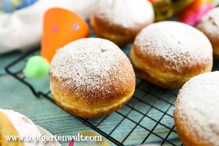 Frisch gebackene Krapfen auf einem schwarzen Abkühlgitter, bestreut mit Staubzucker. Im Hintergrund befinden sich bunte Hütchen.