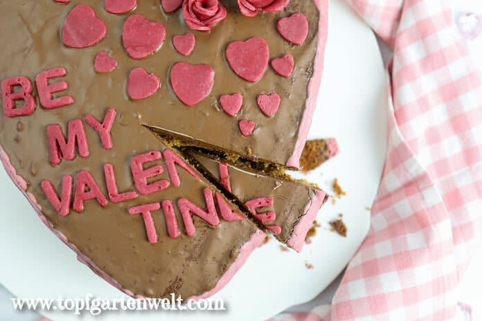 Angeschnittener Kuchen in Herzform. Verziert mit einer Schokoladenglasur. Darauf steht Be my Valentine. Rechts oben sind Rosen und Herzen aus Marzipan.