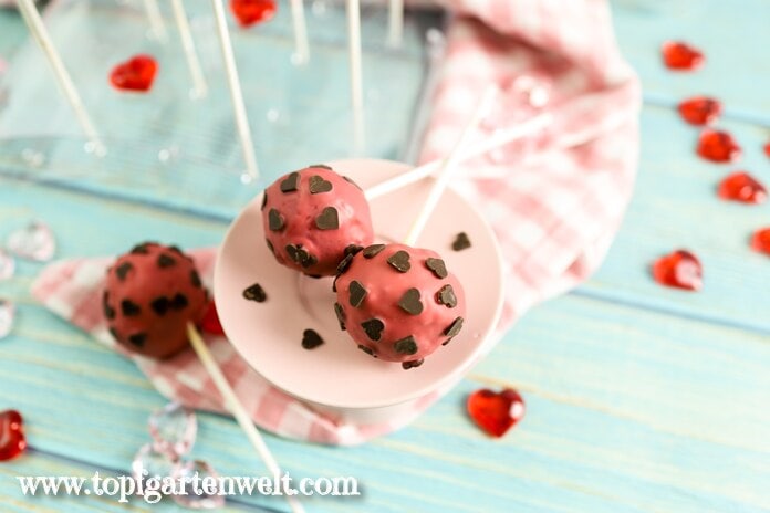 Zwei rosarote Cake Pops verziert mit Schokoherzen liegen auf einer hellrosa Etagere. Im Hintergrund sind rote Herzen als Streudeko zu sehen.