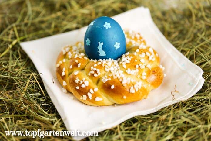 Ein Osterkranz aus Germteig mit einem blauen Ei in der Mitte liegt auf einem weißen Tuch. Als Untergrund dient Heu.