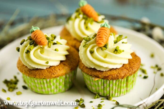 Drei Rübli Muffins in grünen Papierförmchen stehen auf einem weißen Teller. Sie haben ein Frischkäsefrosting, welches mit je einem Marzipan Rübli verziert ist. Darüber sind gehackte Pistazien gestreut.