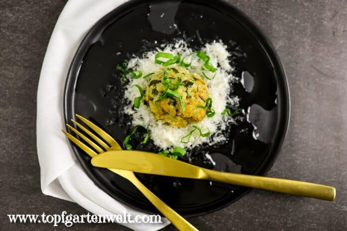 Ein Bärlauchknödel garniert mit fein geriebenen Parmesan und frisch gehackten Bärlauch liegt auf einem schwarzen Teller. Daneben liegt goldenes Besteck.