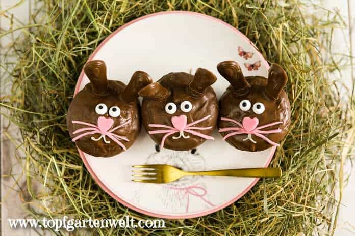 Auf dem Bild sind drei Muffins verziert als Osterhasen zu sehen. Die Ohren sind Katzenzungen, die Augen aus Zucker und Nase und Schnurrbart aus rosa Fondant. Der Mund ist mit Zuckerschrift aufgemalt.