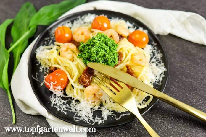 Am Bild ist ein schwarzer Teller mit Spaghetti mit Bärlauchpesto, Tomaten und Garnelen. Darüber ist Parmesan gestreut. Am Rand des Tellers ist goldenes Besteck angelehnt.