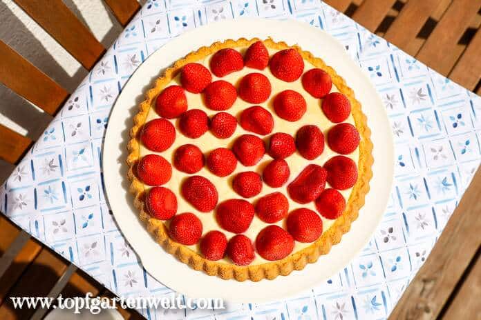 Ein Erdbeerkuchen mit Vanillecreme steht auf einer Gartenbank und ist von oben fotografiert.