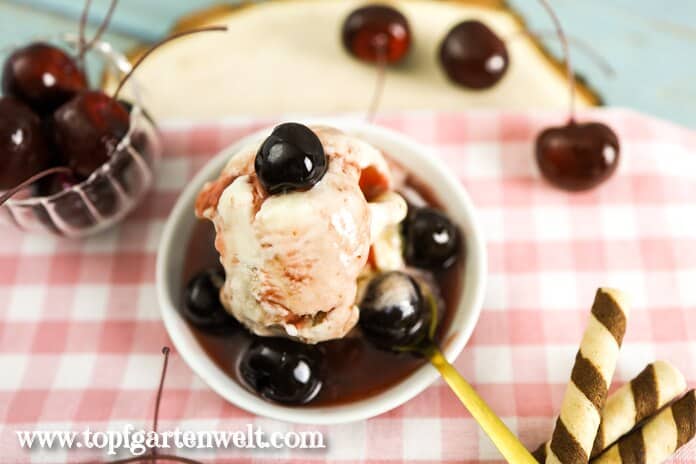 Eine Kugel Amarena Eis ist einem weißen Schälchen angerichtet. Garniert ist sie mit ganz viel Sirup und Früchten. Der Löffel liegt schon bereit.