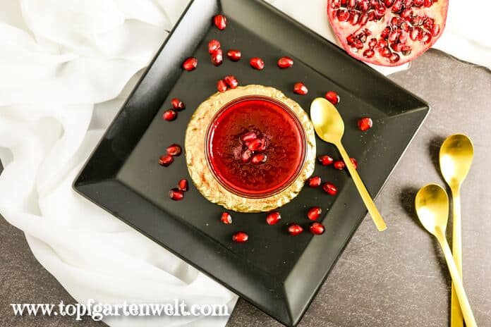 Eine Schale mit Granatapfel Marmelade steht auf einem goldenen Untersetzer, welcher auf einem schwarzen Teller platziert ist. Daneben liegt ein goldener Löffel. Eine Frucht und einige Kerne sind im Hintergrund zu sehen.