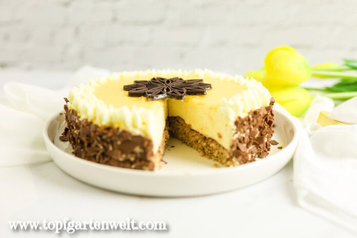 Eine Eierlikörtorte, deren Rand mit Schoko-Flocken verziert ist, steht auf einem weißen Teller. Im Hintergrund liegt eine gelbe Tulpe.