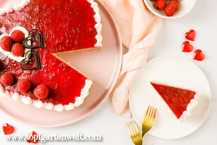 Eine Himbeertorte in Herzform steht auf einem rosaroten Teller. Sie ist angeschnitten. Ein Stück der Torte ist auf einem weißen Teller. Daneben liegt eine goldene Gabel. Als Streudeko sind rote Herzen zu sehen.